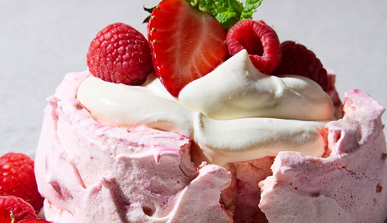 A close-up of a pink meringue dessert topped with whipped cream, fresh raspberries, and a slice of strawberry, garnished with mint.