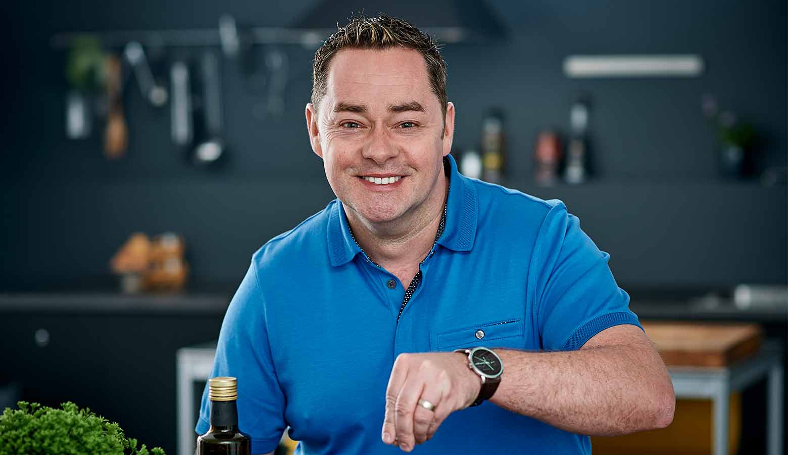 Neven Maguire in a blue shirt sprinkles seasoning in a kitchen. Foreground includes a bottle and greens, with kitchen utensils blurred in the background.