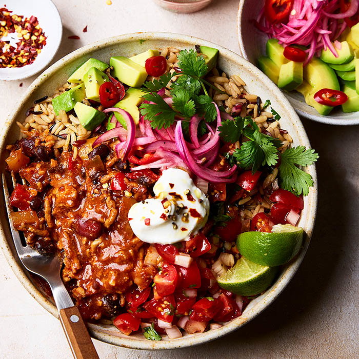 Bowl with rice, chilli, diced avocado, pickled red onions, cherry tomatoes, cilantro, lime wedges, and sour cream topped with chilli flakes.