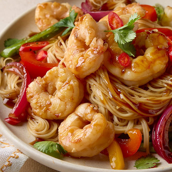 Plate of shrimp stir-fry with noodles, red and yellow bell peppers, red onions, sliced chili, and cilantro garnish.