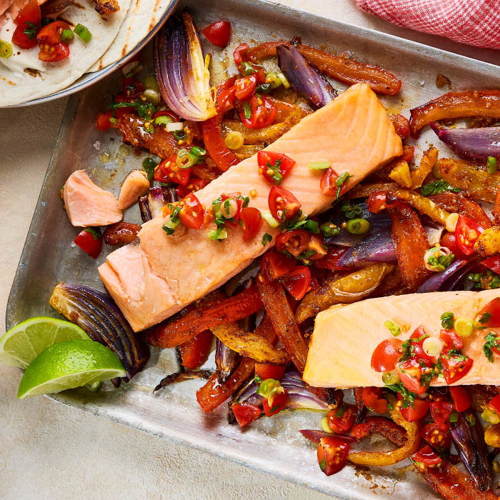 Pan-seared salmon fillets topped with fresh tomato salsa, served on a baking sheet with roasted vegetables and lime wedges.