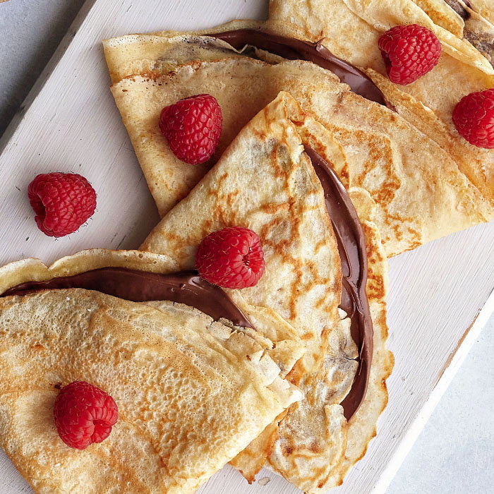 Delicious crepes filled with chocolate spread, garnished with fresh raspberries, arranged on a light wood board.