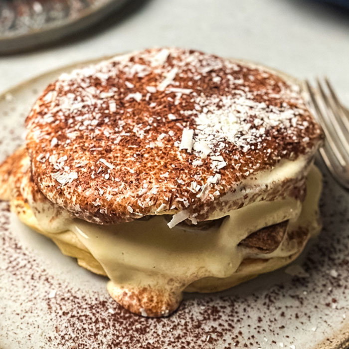 A close-up of two fluffy pancakes layered with creamy custard, topped with coconut flakes and dusted with cocoa powder.