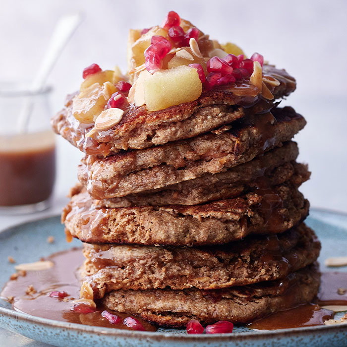 A tall stack of fluffy pancakes topped with caramel sauce, diced apples, pomegranate seeds, and almonds on a blue plate.