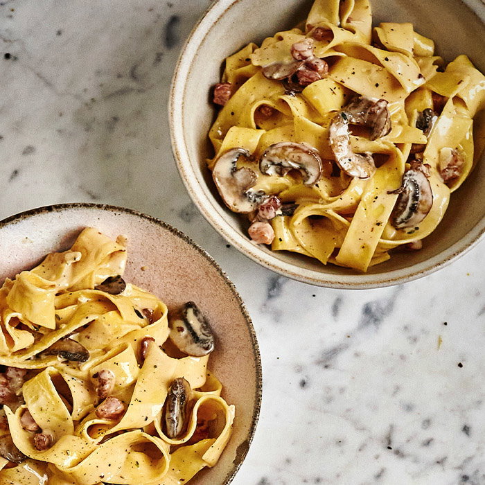 Two bowls of creamy tagliatelle pasta with mushrooms and bacon, sprinkled with black pepper, on a marble surface. The dish looks rich and savory.