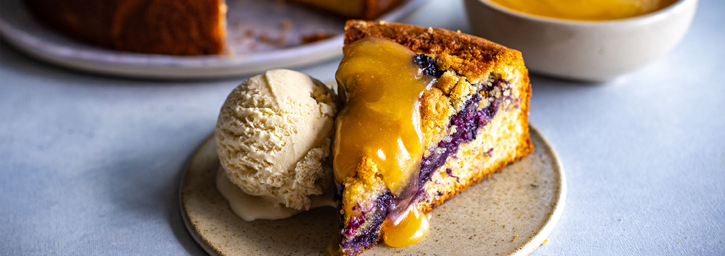 Slice of blueberry cake topped with lemon curd, served with a scoop of vanilla ice cream on a ceramic plate.