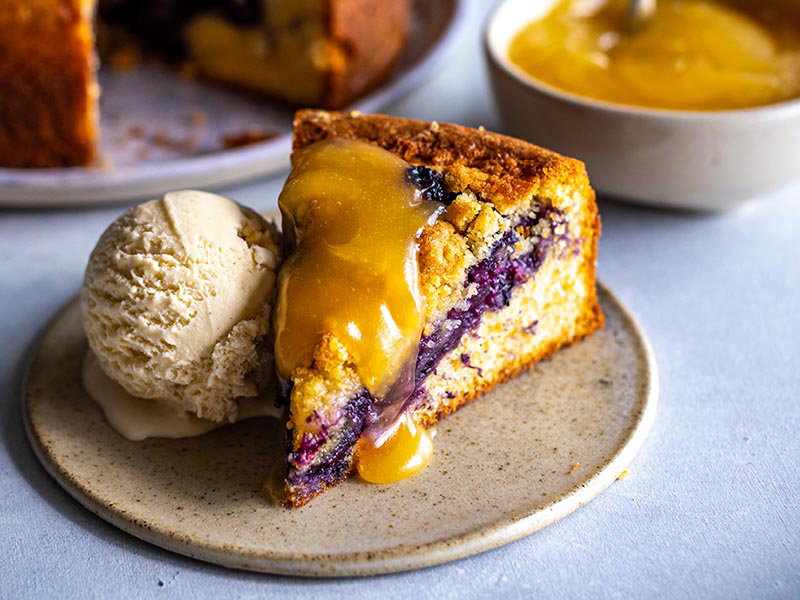 Slice of blueberry cake topped with warm lemon curd, served with a scoop of vanilla ice cream on a ceramic plate.