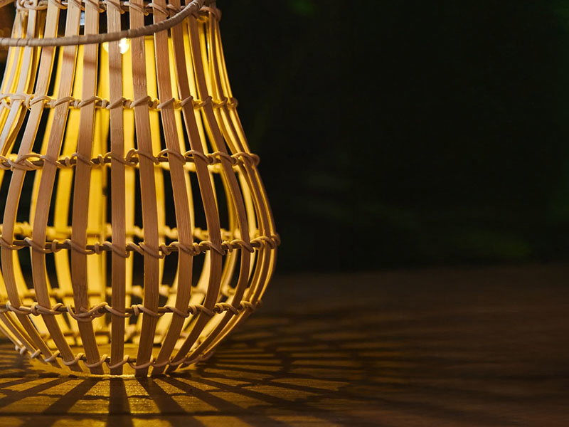 Woven bamboo lantern glowing warmly, casting intricate shadows on a dark wooden surface against a black background.