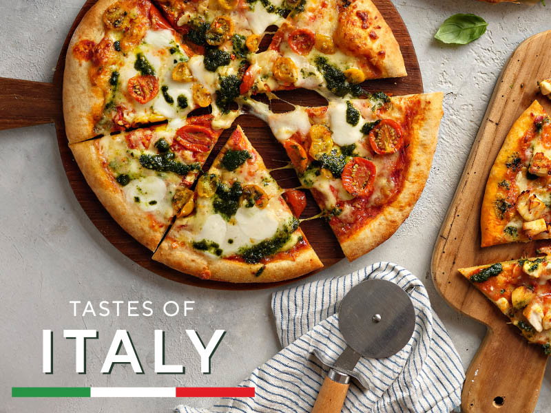 A sliced pizza with cherry tomatoes, cheese, and green pesto sauce on a wooden board, with a pizza cutter and towel nearby.