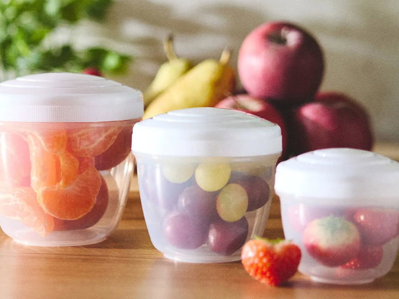 Three clear plastic containers with white lids hold peeled mandarin slices, grapes, and strawberries on a wooden surface with apples and pears in the background.