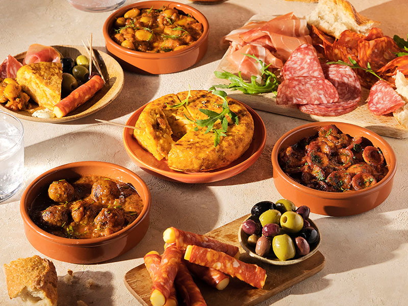 Selection of Spanish tapas including omelette, meatballs, butter beans, chorizo al vino tinto, cured meats and bread, served in rustic terracotta dishes.