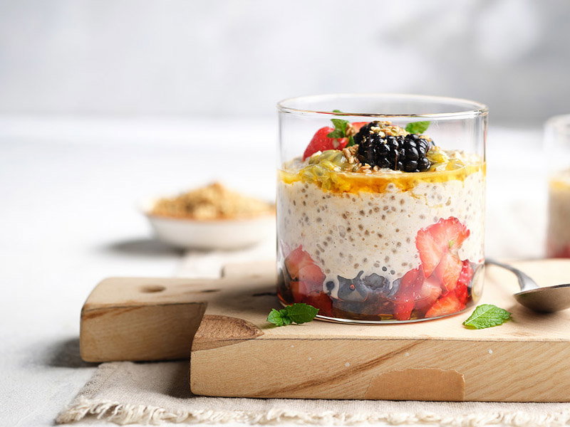 Layered chia pudding parfait in a glass, topped with berries, passion fruit, and mint, sits on a wooden board in a bright kitchen.
