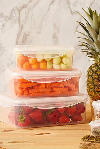Stacked transparent food storage containers filled with strawberries, carrot sticks and melon pieces on a kitchen worktop, with a pineapple in the background.