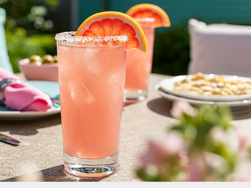 A Paloma cocktail in tall glass with salted rim, garnished with fresh grapefruit slice. Refreshing summer drink photographed on outdoor table setting with additional food items in background.