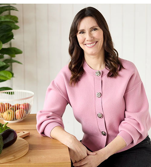 Aileen Cox Blundell is wearing a pink cardigan, sitting in modern kitchen. She's leaning on wooden countertop with hands positioned confidently.