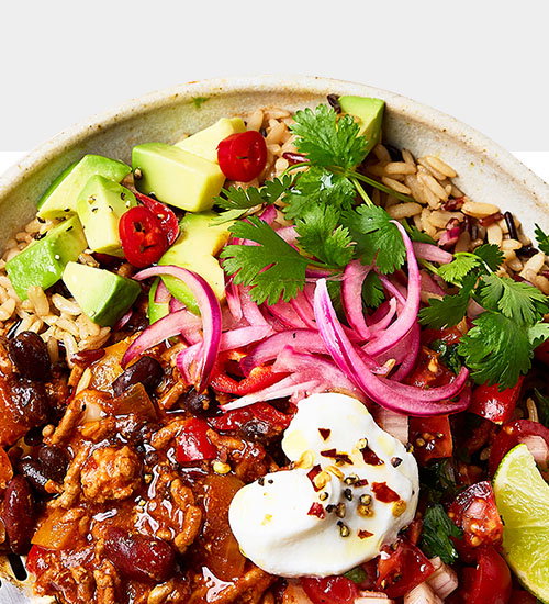 A colourful bowl filled with rice, beans, diced avocado, cherry tomatoes, red onions, cilantro, seasoned meat, and a dollop of sour cream.