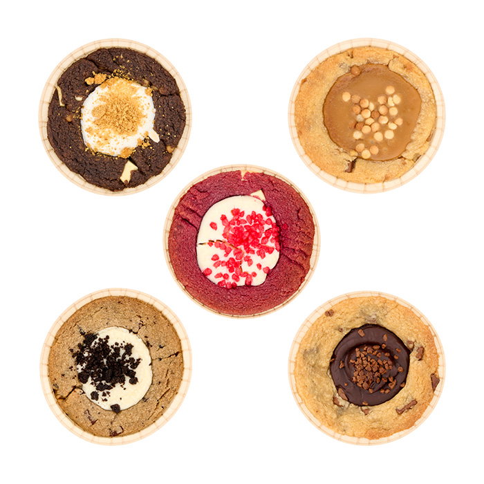Five assorted cookie cups viewed from above, featuring Rocky Road, Chocolate Caramel, Red Velvet, Cookies n&rsquo; Cream and Milk Chocolate Chunk varieties, each with a distinctive filled centre and topping, arranged on a light grey background.