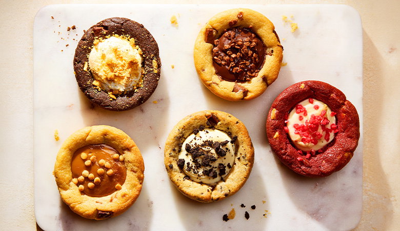 Five assorted cookie cups on a marble serving board, including Rocky Road, Chocolate Caramel, Cookies n&rsquo; Cream, Red Velvet and Milk Chocolate Chunk flavours, viewed from above.