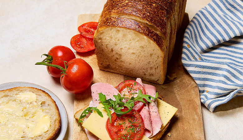 Sliced sourdough white pan bread loaf on a wooden board with ham, cheese and tomato sandwich ingredients and a buttered slice