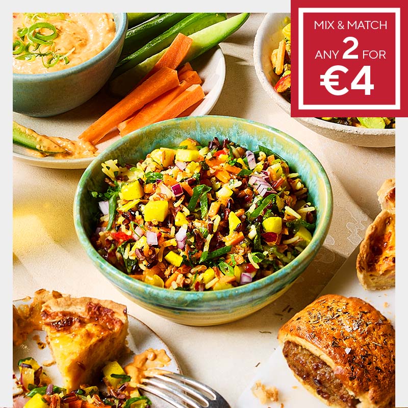 Colorful salad in a bowl surrounded by veggie sticks, quiche, and a pastry roll. A red sign offers "Mix & Match Any 2 for €4," adding a promotional tone.