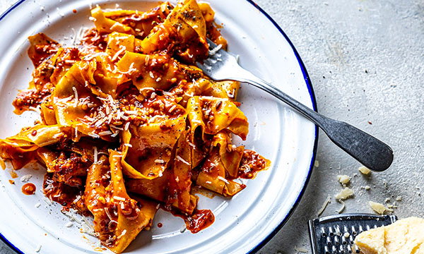 A white plate with chorizo carbonara pasta coated in rich tomato sauce, sprinkled with Parmesan. A fork rests on the side. Cheese and a grater are nearby on the table.