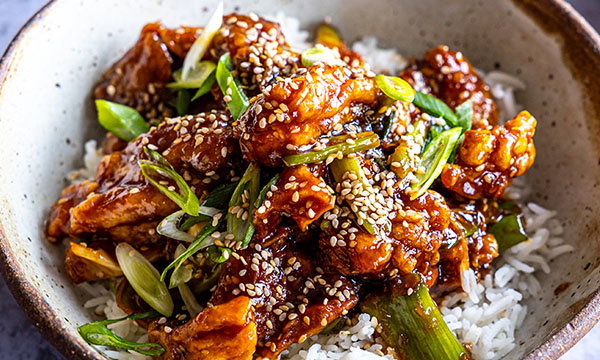 A bowl of savoury sesame chicken topped with sliced green onions and sesame seeds, served on a bed of white rice, conveying a delicious, homemade feel.