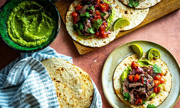 Carne asada tacos served on soft tortillas and topped with fresh salsa, coriander and lime wedges on a rustic platter.
