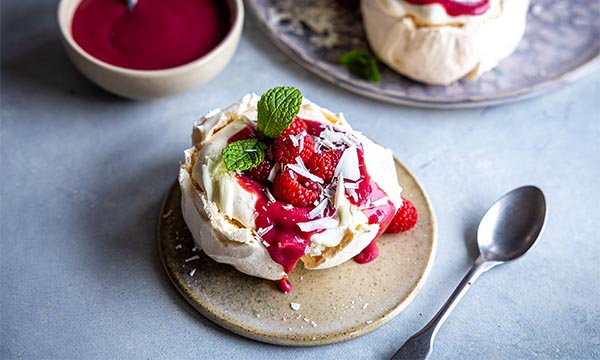A pavlova topped with whipped cream, fresh berries, and drizzled with raspberry sauce. Garnished with mint leaves, placed on a rustic plate beside a spoon.