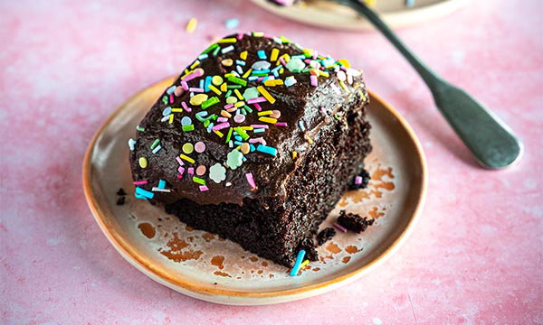 A slice of chocolate cake with glossy icing and colourful sprinkles sits on a beige plate. The background is a soft pink surface, and a fork is nearby.