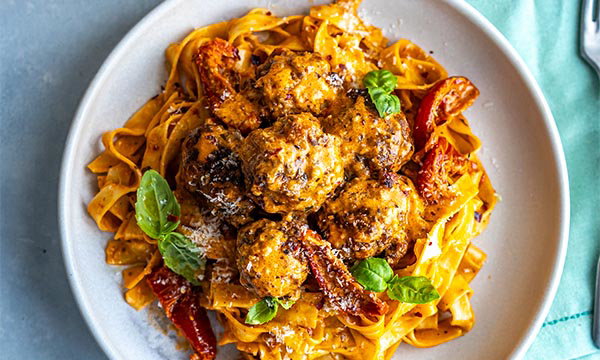A plate of pasta with creamy meatballs, sun-dried tomatoes, and fresh basil, on a light blue table. The dish looks rich and appetising.