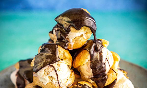 Cream puffs stacked on a plate, filled with vanilla ice cream and drizzled with glossy chocolate sauce, set against a blurred blue and green background.