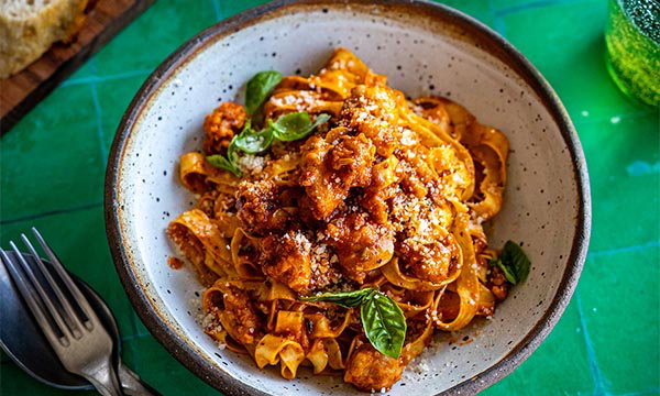 Bowl of tagliatelle pasta with rich tomato sauce, topped with fresh basil and grated cheese, set on a vibrant green table. Fork and spoon beside it.