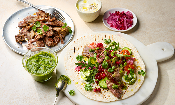 Grilled tortilla topped with lamb, coriander, cherry tomatoes, cucumbers, red onions, and green sauce. Plates nearby hold extra lamb, pickled onions, and sauce.