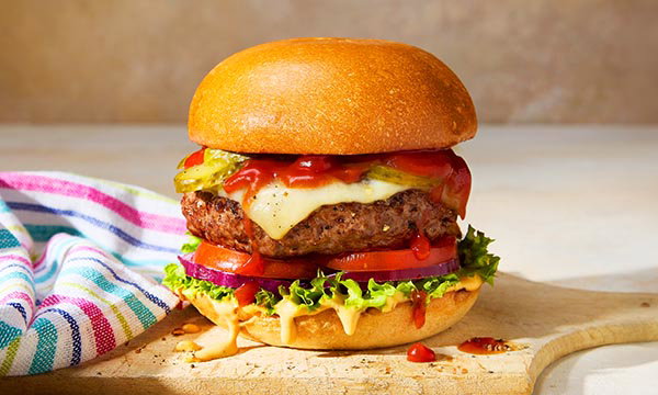 A juicy cheeseburger with lettuce, tomato, pickles, and sauce, on a wooden board. A colourful striped cloth is beside it, creating a fresh, appetising look.