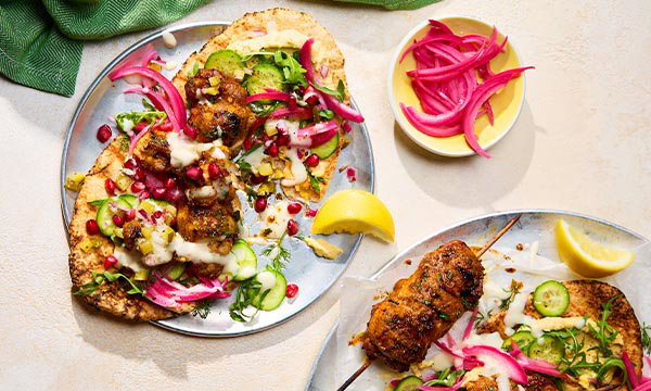 Chicken shawarma flatbreads topped with pickled red onions, cucumber, herbs, pomegranate seeds and creamy sauce, served with lemon wedges and a side of pickled onions.