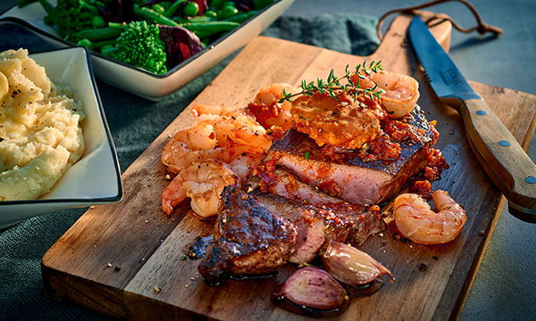 Grilled steak topped with prawns and herbs on a wooden board, accompanied by a knife. Sides include mashed potatoes and saut&eacute;ed green vegetables.