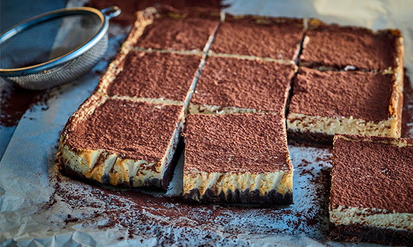 Nine chocolate-covered cheesecake squares on parchment paper, with a sifter to the left. The image conveys a rustic and inviting dessert scene.