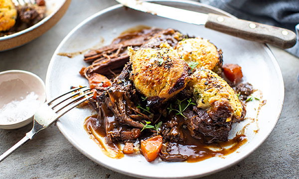 Slow-cooked beef with golden-brown biscuits, carrots, and rich gravy on a white plate. Fork and knife beside the dish showcasing a hearty meal.