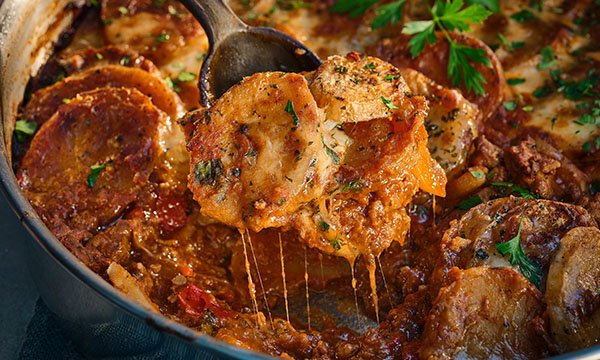 A close-up of a rich, golden-brown potato casserole in a pan, garnished with herbs. A spoon lifts a cheesy, well-baked slice, evoking warmth and comfort.