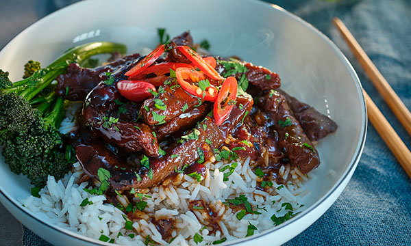 A white bowl filled with savoury beef in a glossy sauce atop fluffy white rice, garnished with sliced red chillies and green herbs. Tenderstem broccoli is on the side. Chopsticks rest nearby, creating a fresh, appetising presentation.