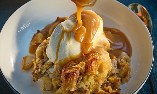 A warm bread pudding topped with a scoop of vanilla ice cream is drizzled with rich caramel sauce in a white bowl, creating an indulgent and cozy dessert scene.