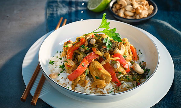 A vibrant Thai curry dish with chicken, red peppers, and greens atop white rice, garnished with parsley. A lime wedge and cashews are in the background.