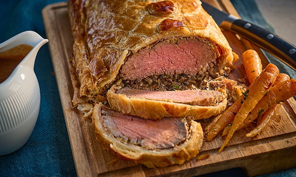 A golden-brown Beef Wellington on a wooden board, sliced to reveal pink, juicy beef and mushroom filling, served with roasted carrots and gravy.