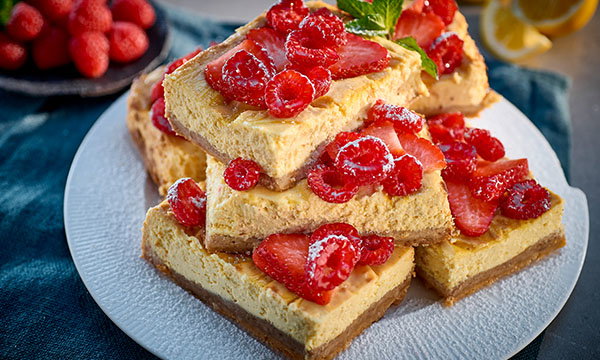 A stack of creamy cheesecake bars topped with fresh raspberries and sliced strawberries, garnished with mint leaves on a white plate. A vibrant, appetizing dessert display.