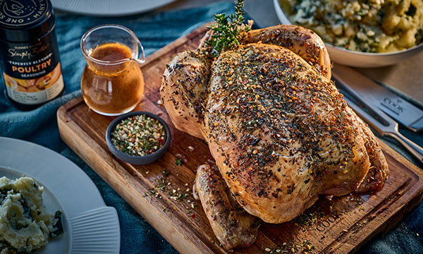 Roasted chicken seasoned with herbs on a wooden board, surrounded by spices, gravy, and mashed potatoes. Warm, inviting culinary setting.