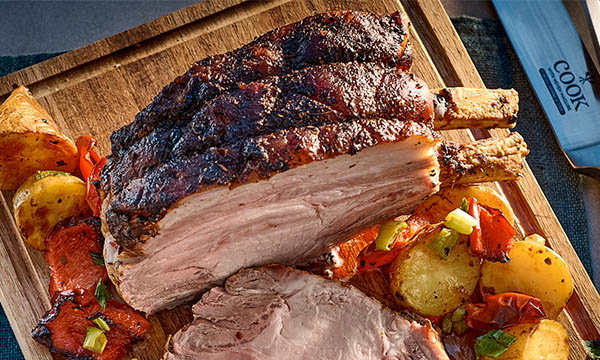 Roasted pork rib on a wooden board with crispy skin, surrounded by golden potatoes and roasted vegetables, next to a chef's knife. Inviting and hearty.