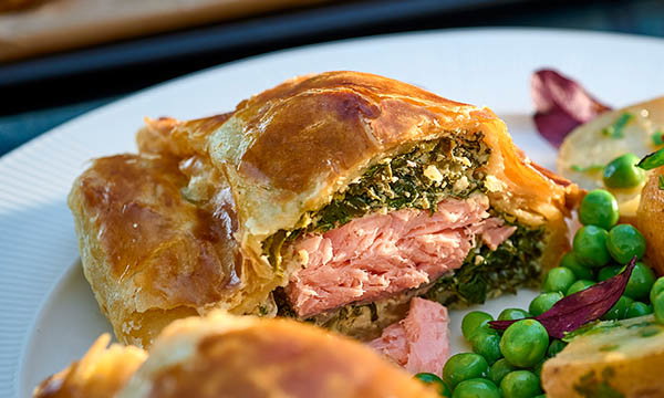 A slice of salmon en croute with flaky golden pastry, filled with salmon and spinach. Served with peas and potatoes, creating a fresh, appetising dish.
