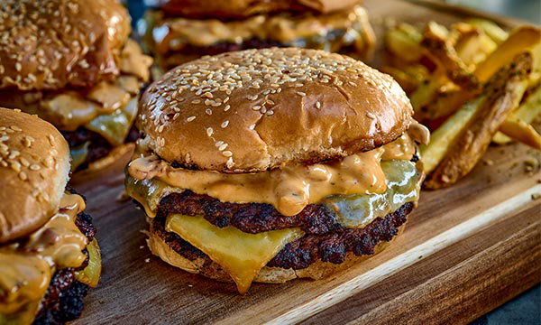 Crispy smash burger with two beef patties, melted cheese, and creamy sauce in a sesame bun. Served with golden fries on a wooden board. Indulgent and savoury.
