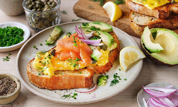 Toasted bread with scrambled eggs, smoked salmon, avocado slices, red onion, and herbs on a plate, garnished with lemon wedges, evokes a fresh brunch vibe.