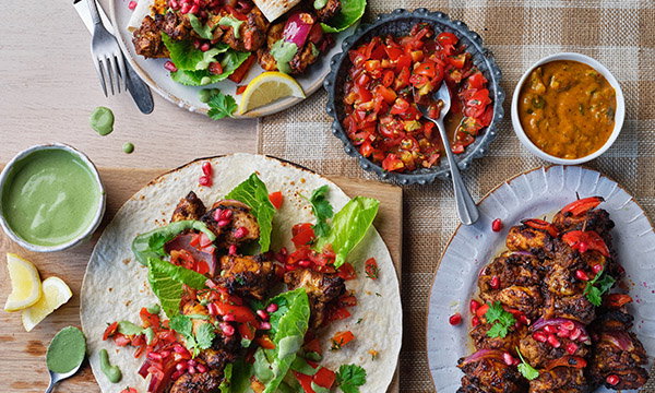 A vibrant spread of tandoori chicken dishes featuring grilled chicken wraps with lettuce and pomegranate seeds. Accompanied by bowls of salsa and curry.